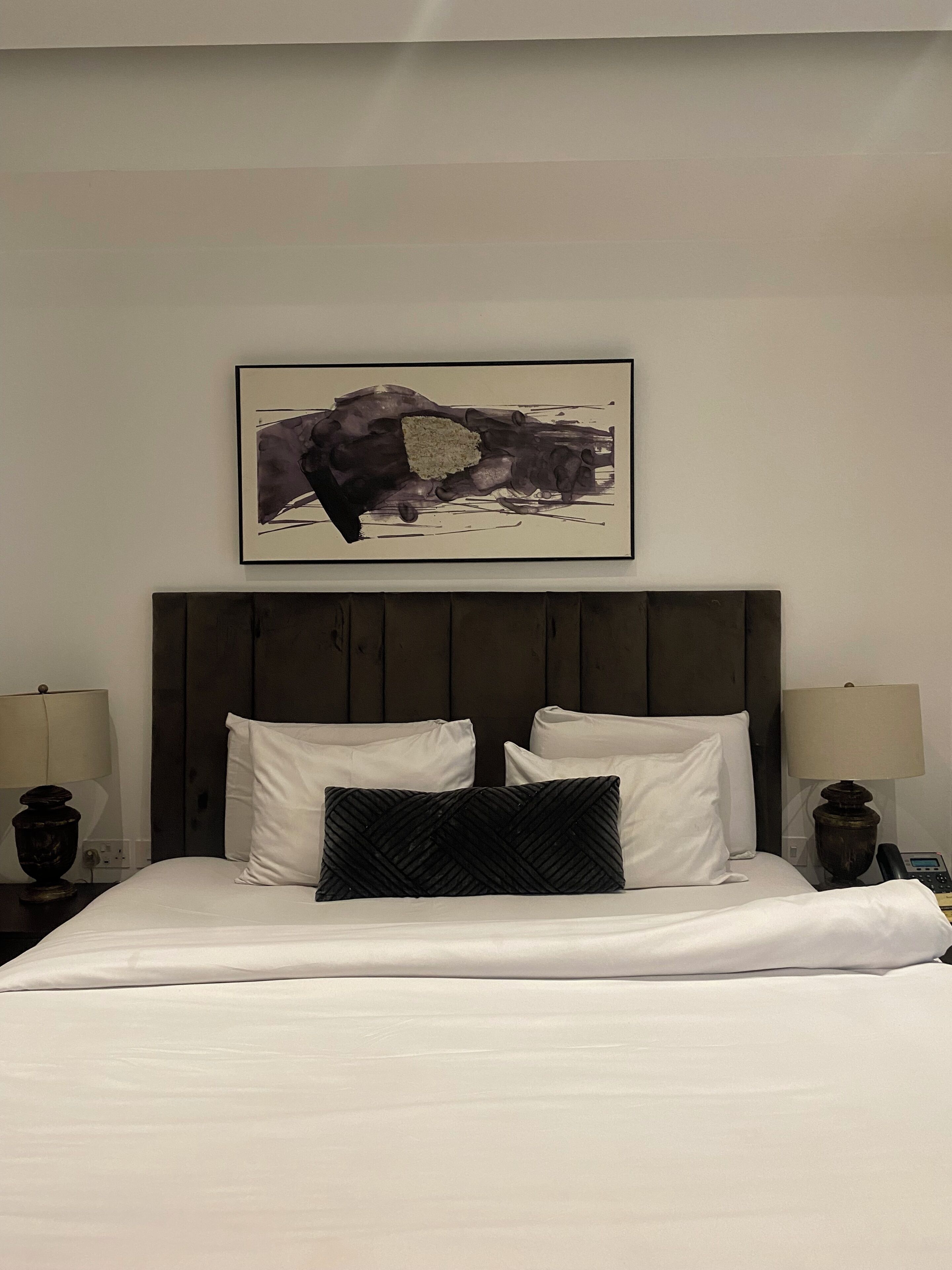 Quarto de hotel com uma cama grande, duas luminárias laterais, uma cabeceira de madeira escura e uma obra de arte abstrata emoldurada sobre a cama.