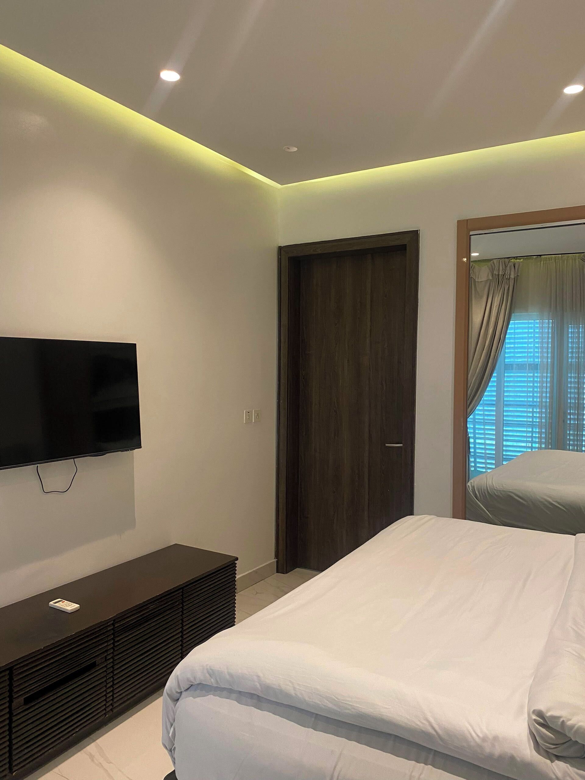 A modern bedroom with a flat-screen TV, a dark wooden wardrobe, a bed with white bedding, and a window with curtains.