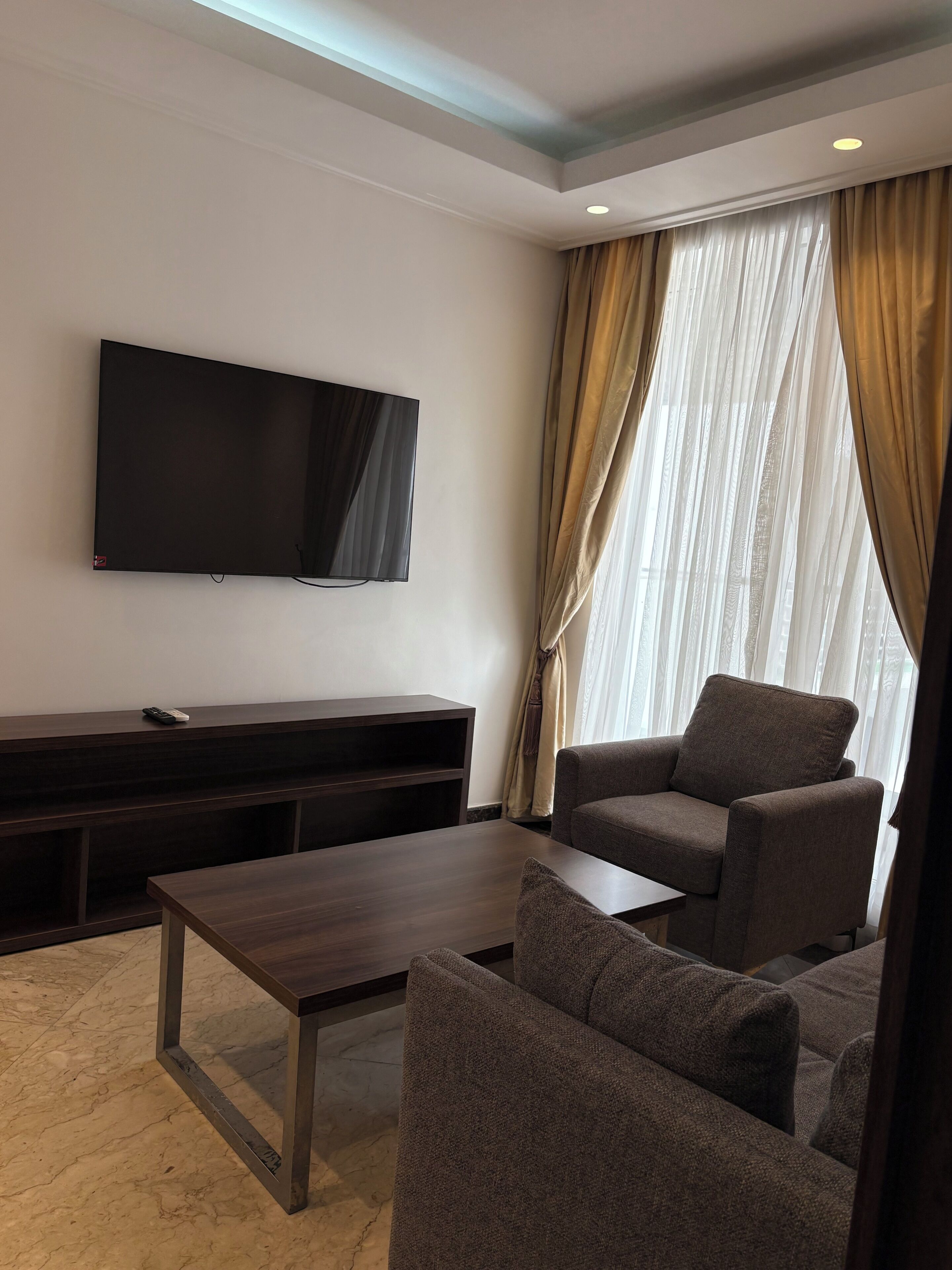 Una sala de estar moderna con un televisor de pantalla plana, muebles de madera oscura y un ventanal con cortinas translúcidas.