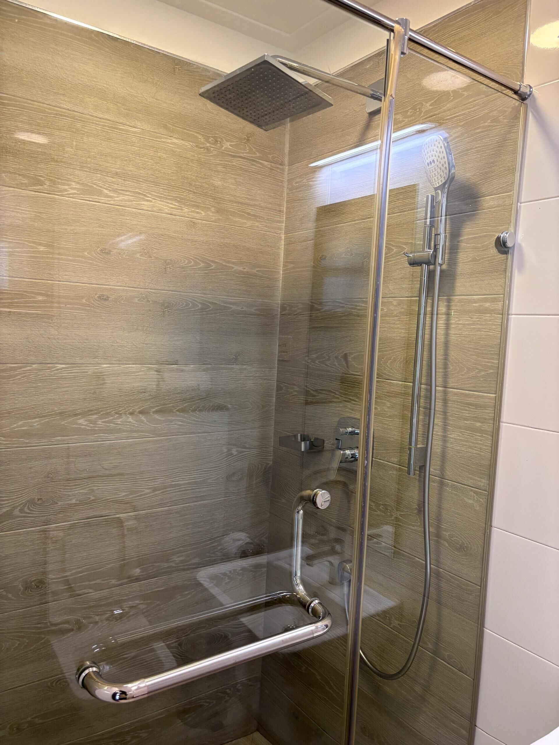 A glass-enclosed shower with a wooden panel wall, a handheld showerhead, and a handheld shower control.