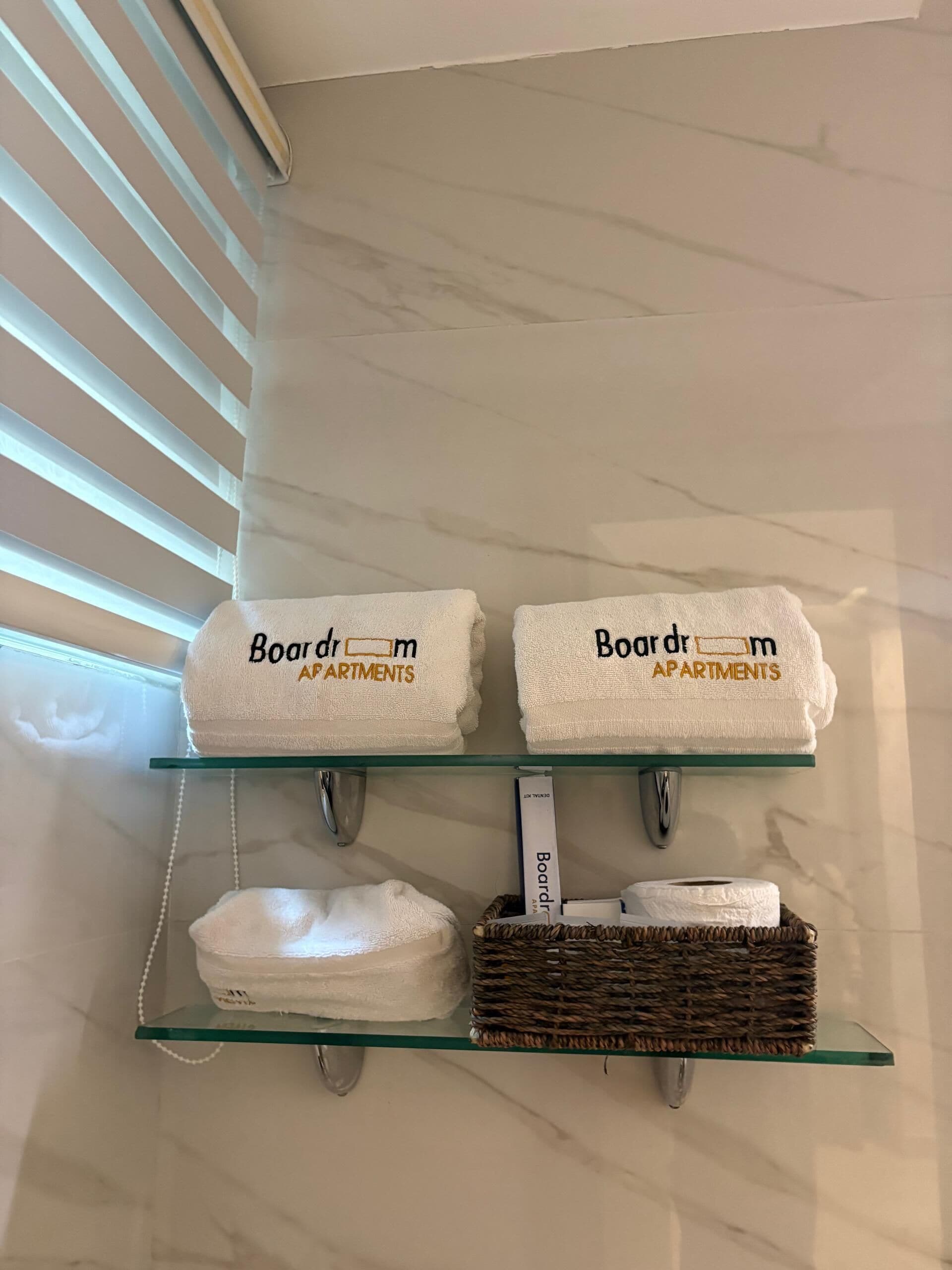 A bathroom with a glass shelf holding two towels labeled "Boardroom Apartments" and a wicker basket.