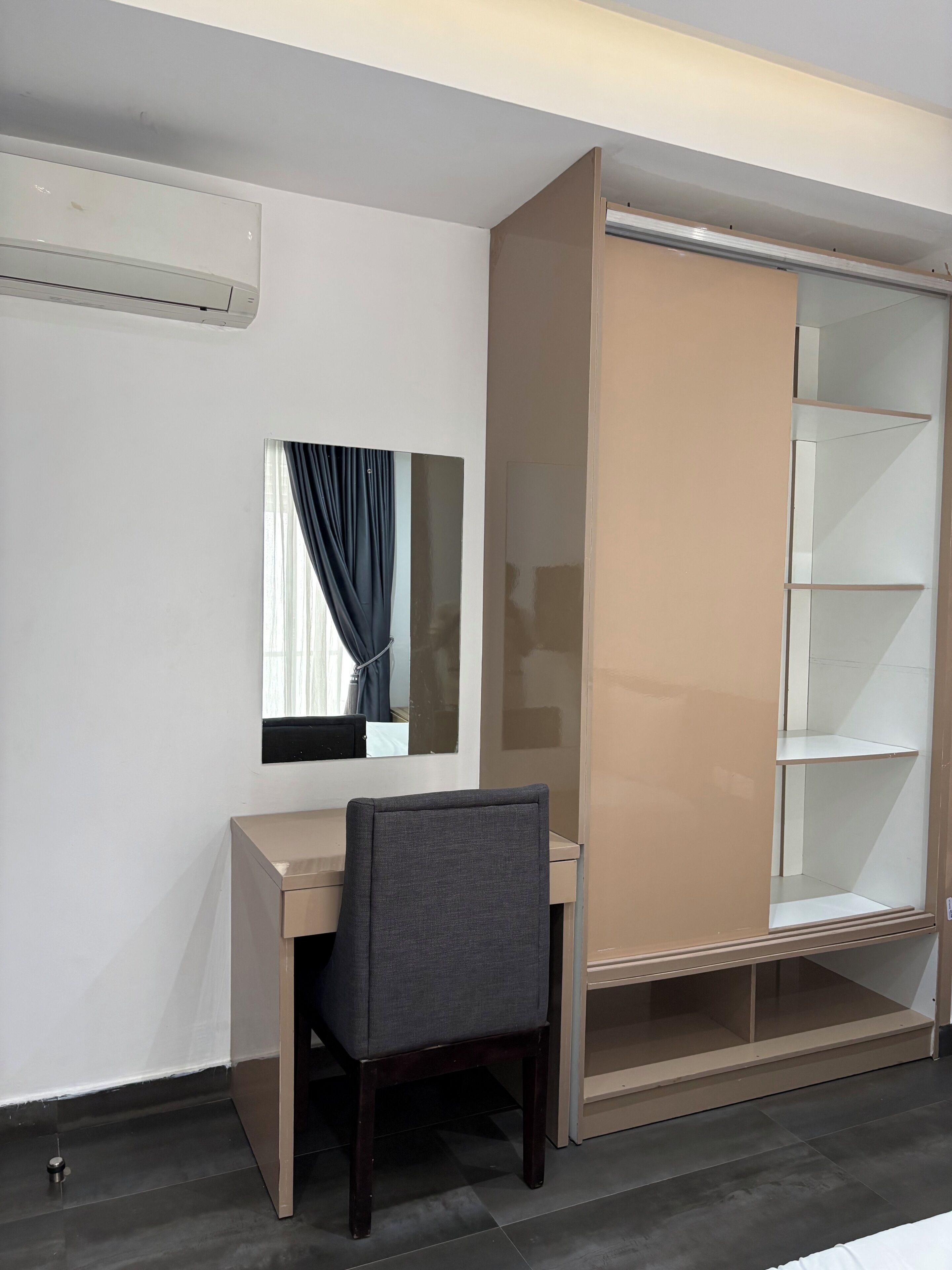 A room with a desk, chair, wardrobe, mirror, and an air conditioning unit.