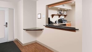 A hotel room with a wooden floor, a black doormat, a white door, and a mirror reflecting a kitchenette area.
