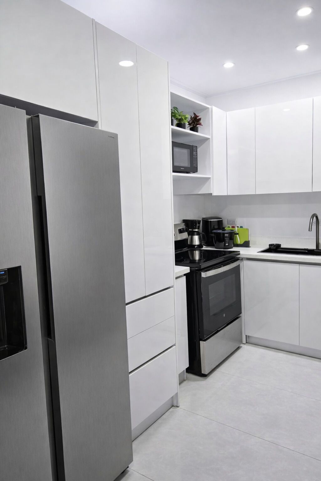 Une cuisine moderne avec des meubles blancs, un réfrigérateur en acier inoxydable, un four noir et un micro-ondes posé sur une étagère.