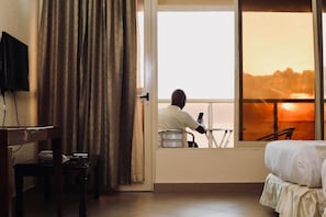 A hotel room with a bed, a desk, a chair, a television, and a window with a view of the sunset. - The Landmark Mbezi Beach Resort and Conference Centre (Dar es Salaam)