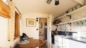 A compact kitchen with white cabinets, a wooden dining table, and a view into the adjacent bathroom. - Plank Bridge | Shepherds Hut in Apple Orchard | Sleeps 2 (West Ayton)