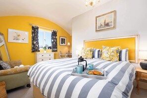 Ein Schlafzimmer mit einem Bett, einem Nachttisch, einem Sessel, einem Fenster mit Vorhängen, einem Gemälde an der Wand und einem Tablett mit Teekanne und Gebäck auf dem Bett.