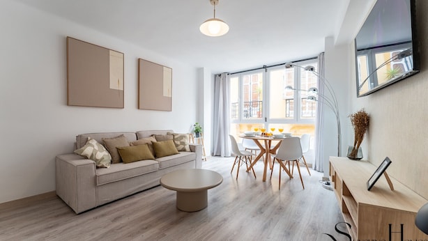 Un salon moderne comprenant un canapé, une table basse et un coin repas. De grandes fenêtres laissent entrer la lumière naturelle et offrent une vue sur l’extérieur.