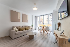 Una sala de estar moderna con sofá, mesa de centro y comedor. Amplias ventanas ofrecen luz natural y vistas al exterior.