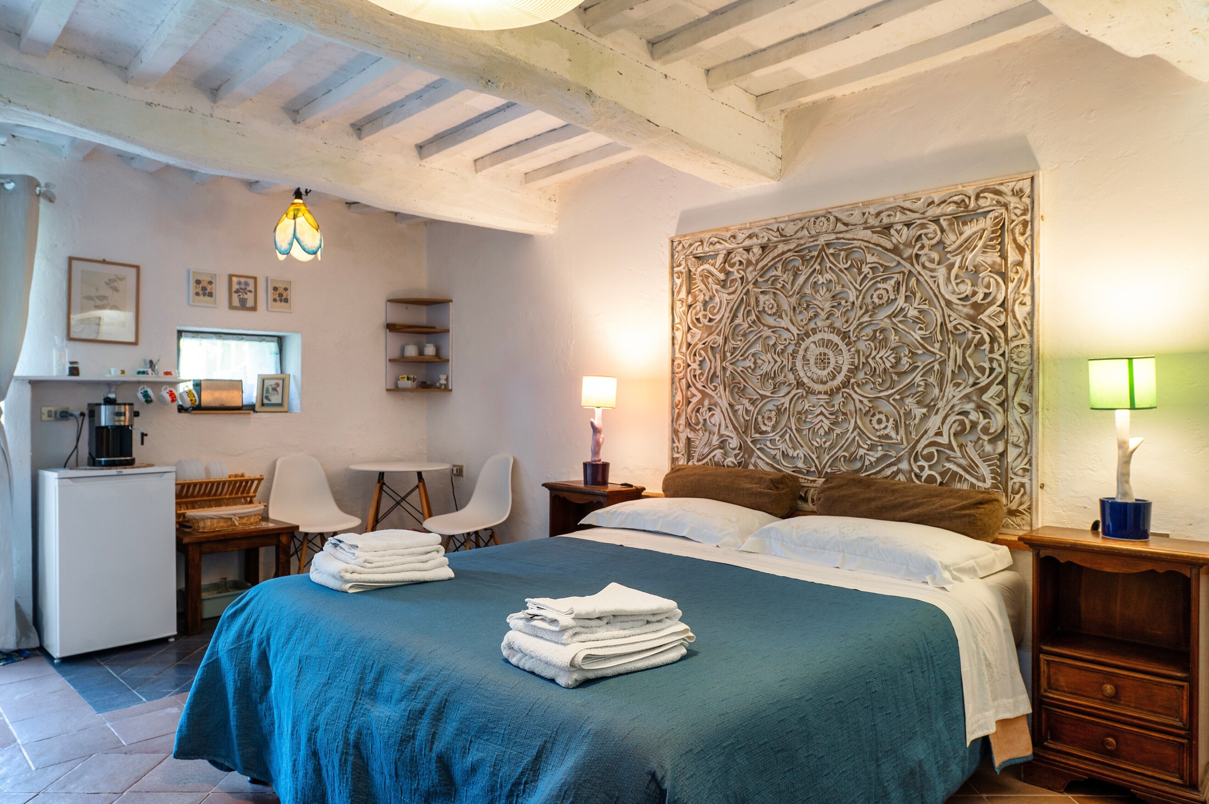 A bedroom with a bed, bedside tables, a desk, chairs, a coffee machine, and a decorative headboard.
