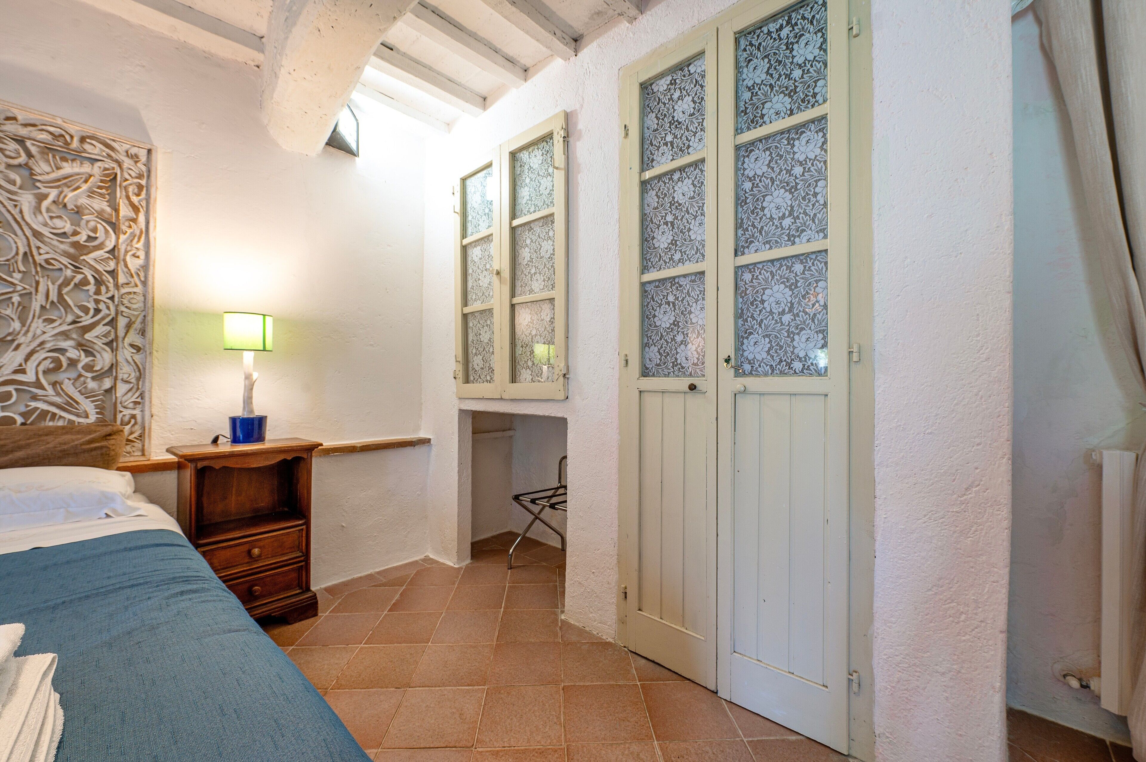 Ein Schlafzimmer mit einem Bett, einem Nachttisch, einer Lampe, einem Kleiderschrank und einem Fenster mit gemustertem Glas.