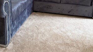 Un sofá modular con tela oscura y costuras decorativas, ubicado sobre una alfombra beige en una habitación con una pared lisa y un papel tapiz texturizado en un lateral.