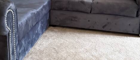 A sectional sofa with a dark fabric and decorative stitching, positioned on a beige carpet in a room with a plain wall and a textured wallpaper on one side.