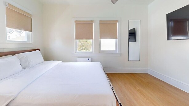 Habitación de hotel con una cama grande, dos ventanas con persianas, un televisor montado en la pared y piso de madera.
