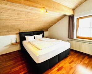 Un dormitorio con techo de madera, una cama grande, una mesita de noche, una ventana con cortinas y un radiador.