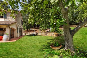 Un bâtiment en pierre avec un toit en tuiles, une cour verdoyante et un arbre orné d’un grand pot de fleurs.
