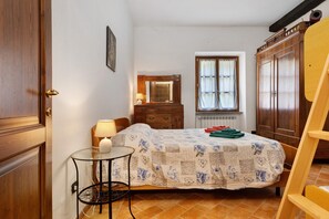 Une chambre à coucher comprenant un lit, une commode, un miroir, une fenêtre avec des rideaux, une petite table avec une lampe et une chaise.