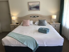 Una cama bien hecha con almohadas, una colcha y toallas. Hay dos mesitas de noche con lámparas. Un cuadro cuelga en la pared sobre la cama.