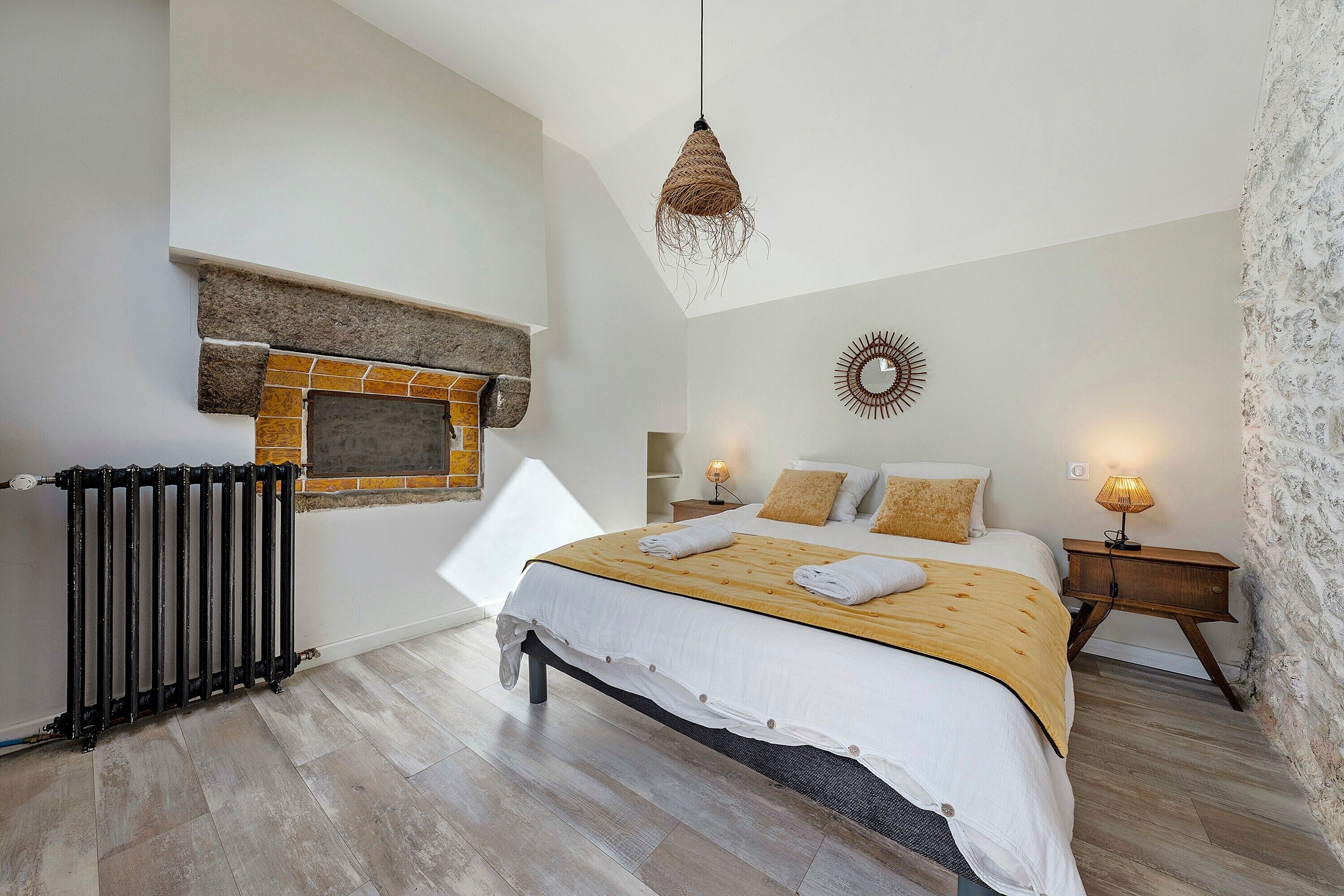 A bedroom with a bed, a fireplace, a hanging light fixture, and a nightstand with a lamp.