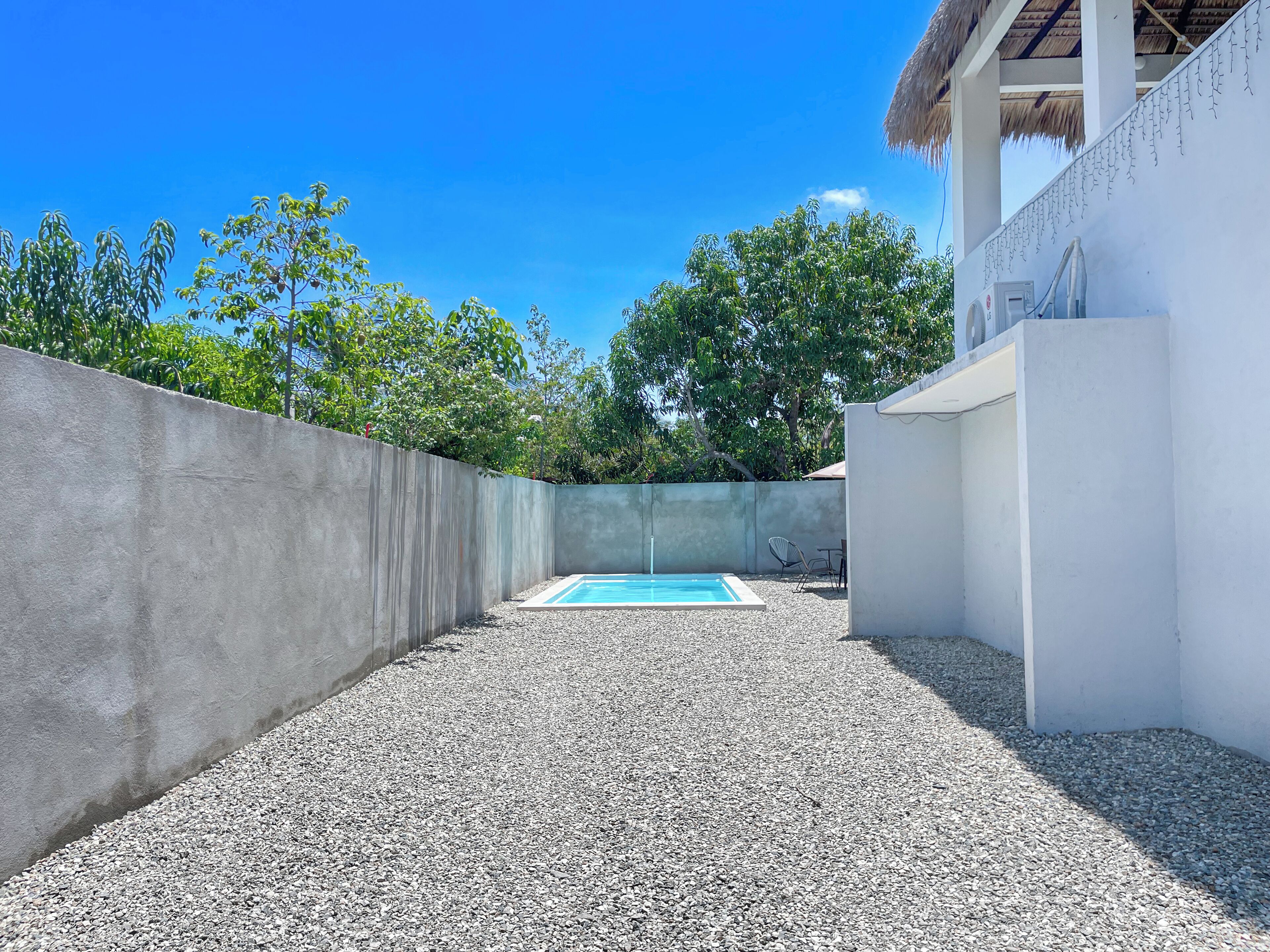 Una piscina amb mur de formigó i sòl de grava, envoltada d'arbres i un edifici blanc.