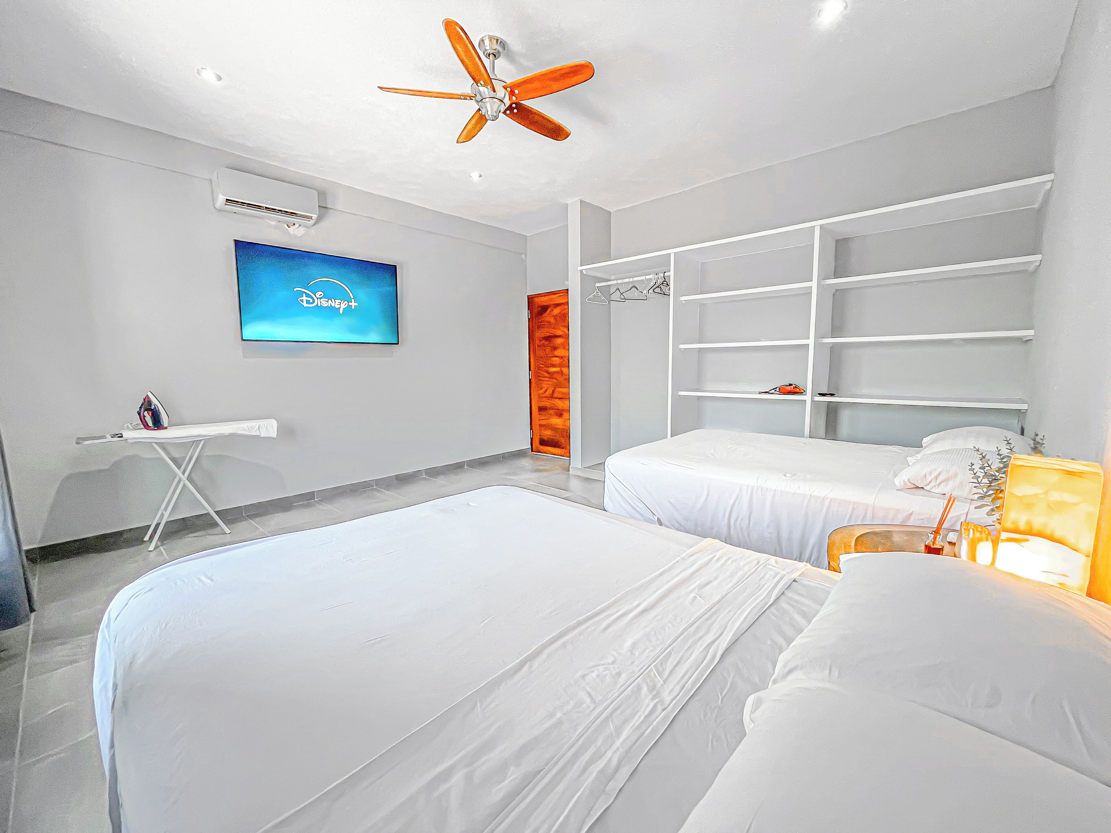 Una habitación de hotel moderna con dos camas, un televisor que muestra el logo de Disney+ y un ventilador de techo, además de un armario con estantes.