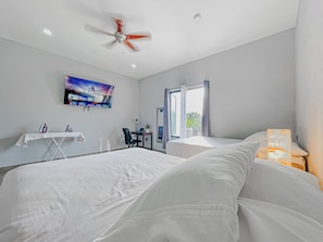 Una camera d'albergo con un letto, una scrivania, una sedia, una ventola a soffitto, una televisione e una finestra con tende.