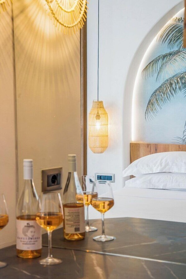 A hotel room with a bed, a wall-mounted painting, a table with wine bottles and glasses, and a hanging light fixture. - Drossia Hotel (Hersonissos)