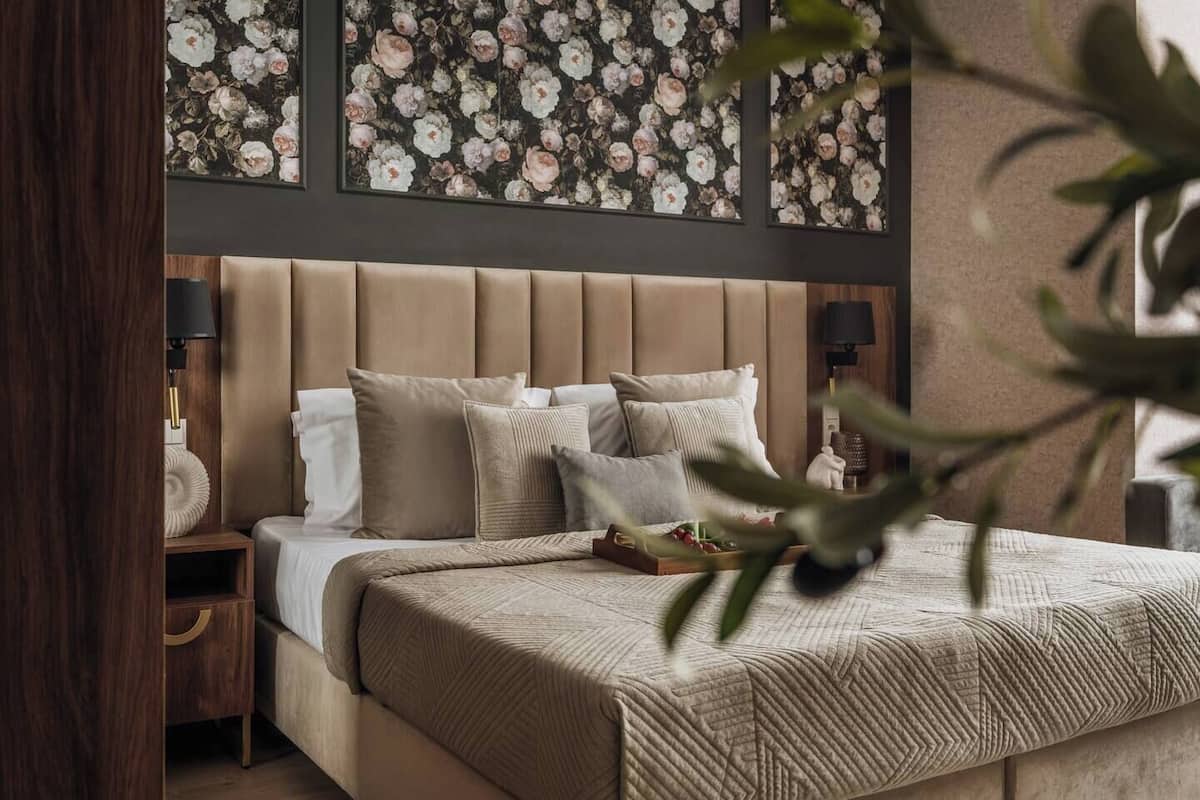 Un dormitorio con cabecera capitonada, cama con múltiples almohadas, mesita de noche con lámpara y una obra de arte floral en la pared.