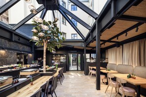 A modern interior with a high ceiling, exposed beams, and a central tree. There are tables and chairs arranged for dining, and a bar area with shelves of bottles. - White Lotus by Fragola Collection (Kraków)