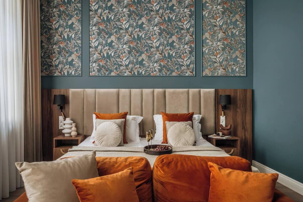 Un dormitorio con cabecera capitonada, dos mesitas de noche con lámparas, y un papel tapiz con motivos florales sobre la cama.