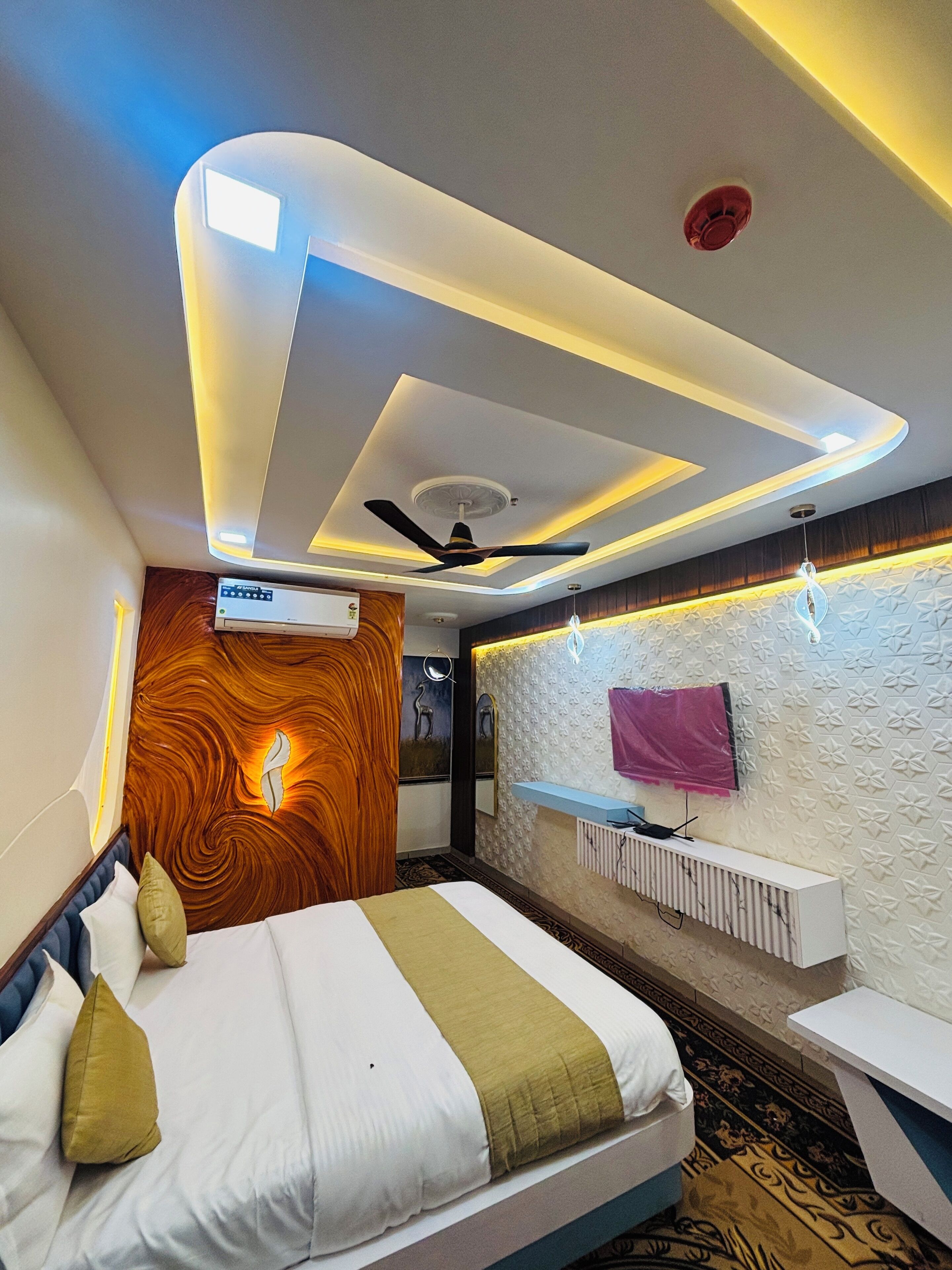 Una habitación de hotel moderna con una cama, ventilador de techo, aire acondicionado montado en la pared, tocador con espejo y un panel decorativo.
