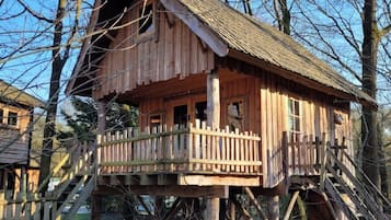 Une cabane en bois perchée dans un arbre, dotée d’un balcon et construite sur pilotis, entourée d’arbres.
