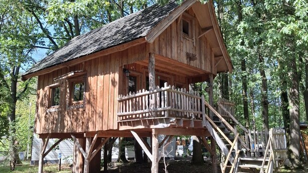Una cabaña de madera con un balcón y una escalera que lleva a él, rodeada de árboles.