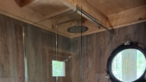 Una ducha envuelta en vidrio con cabezal de ducha manual, una ventana pequeña y un ventanal redondo.