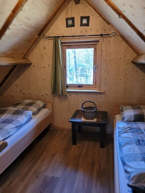 A cozy attic bedroom with two beds, a small table, and a window with a view of trees.