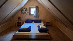 Un dormitorio acogedor y pequeño en el ático, con techo inclinado de madera, una cama individual con ropa de cama azul, dos mesitas de noche y una ventana con cortinas.