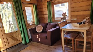 Un interior de cabaña acogedor, con piso de madera, un sofá marrón y un cojín redondo, una mesa de comedor de madera y una ventana con cortina verde.