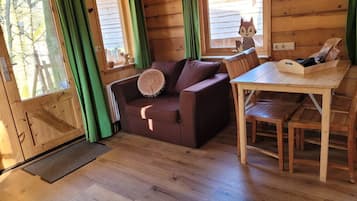 Ein gemütliches Blockhaushaus-Interieur mit Holzboden, einem braunen Sofa mit rundem Kissen, einem Holzesstisch und einem Fenster mit grünem Vorhang.