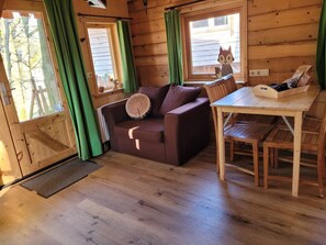 Un interior de cabaña acogedor, con piso de madera, un sofá marrón y un cojín redondo, una mesa de comedor de madera y una ventana con cortina verde.