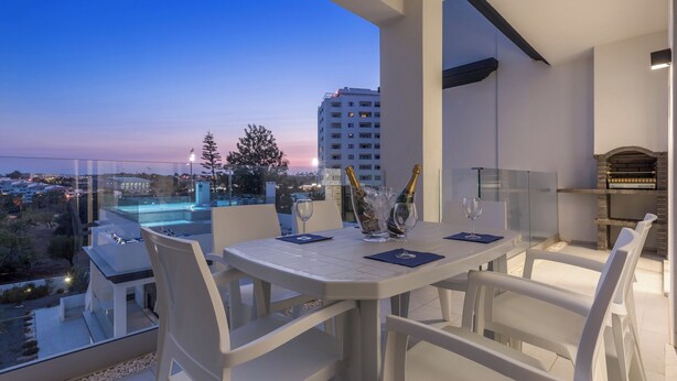 A balcony with a table set for two, a glass partition, and a view of the cityscape at dusk. - Correeira Luxury Residence E (Albufeira)