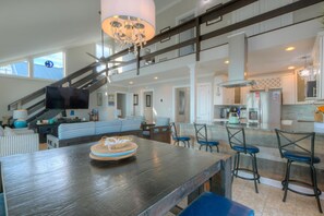Amplia sala de estar con una gran mesa de madera, sillas azules, televisor y una escalera que conduce a un nivel superior.