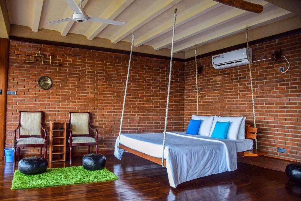 Een kamer met een bakstenen muur, een zwevend bed, twee stoelen, een groene vloerkleed en een plafondventilator.
