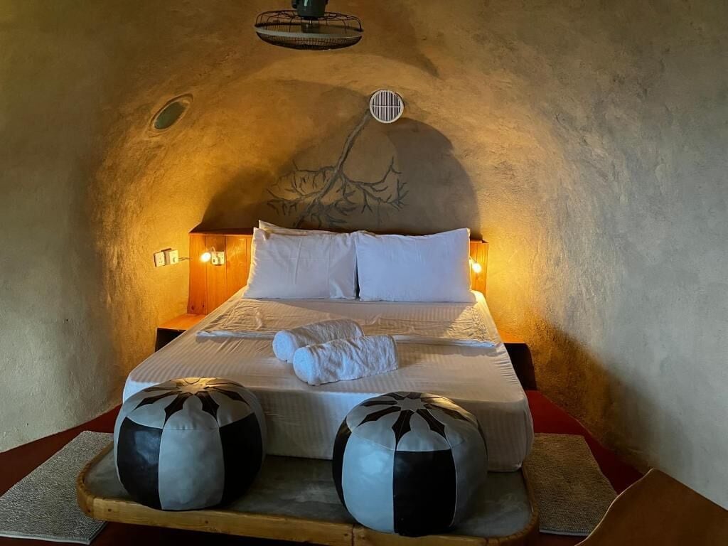 Quarto com teto em arco singular, cama com roupa de cama branca, duas almofadas decorativas e luminária fixada na parede.