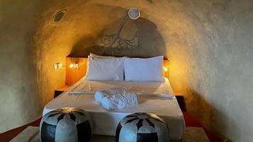 Quarto com teto em arco singular, cama com roupa de cama branca, duas almofadas decorativas e luminária fixada na parede.