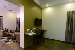 Een knus woongedeelte met een bank, een salontafel, een bureau met een stoel, een flatscreen-tv aan de muur en een raam met gordijnen.