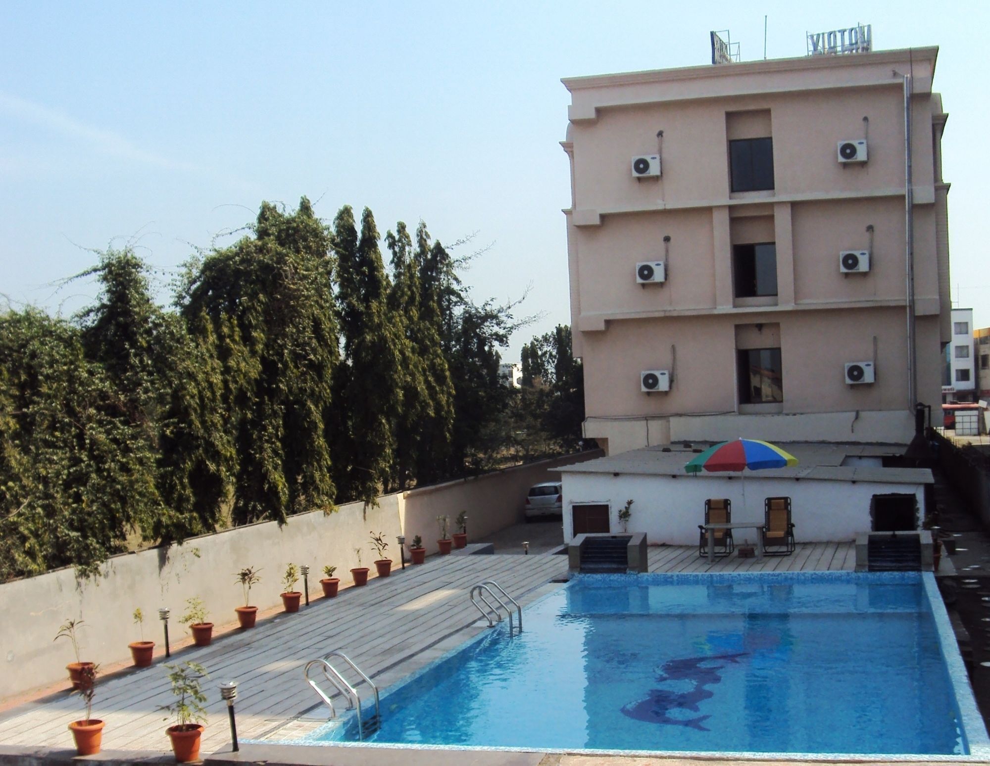 A rooftop swimming pool with a view of a multi-story building, potted plants, and a small seating area.