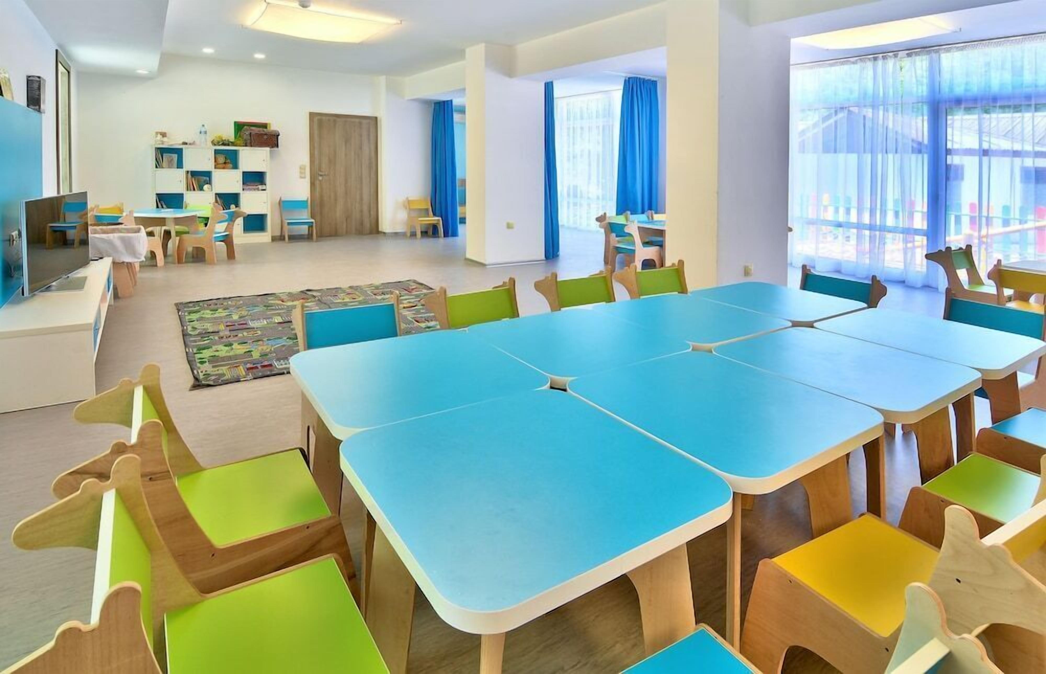 A spacious room with a large blue table and colorful chairs, a bookshelf, and a play area with toys.
