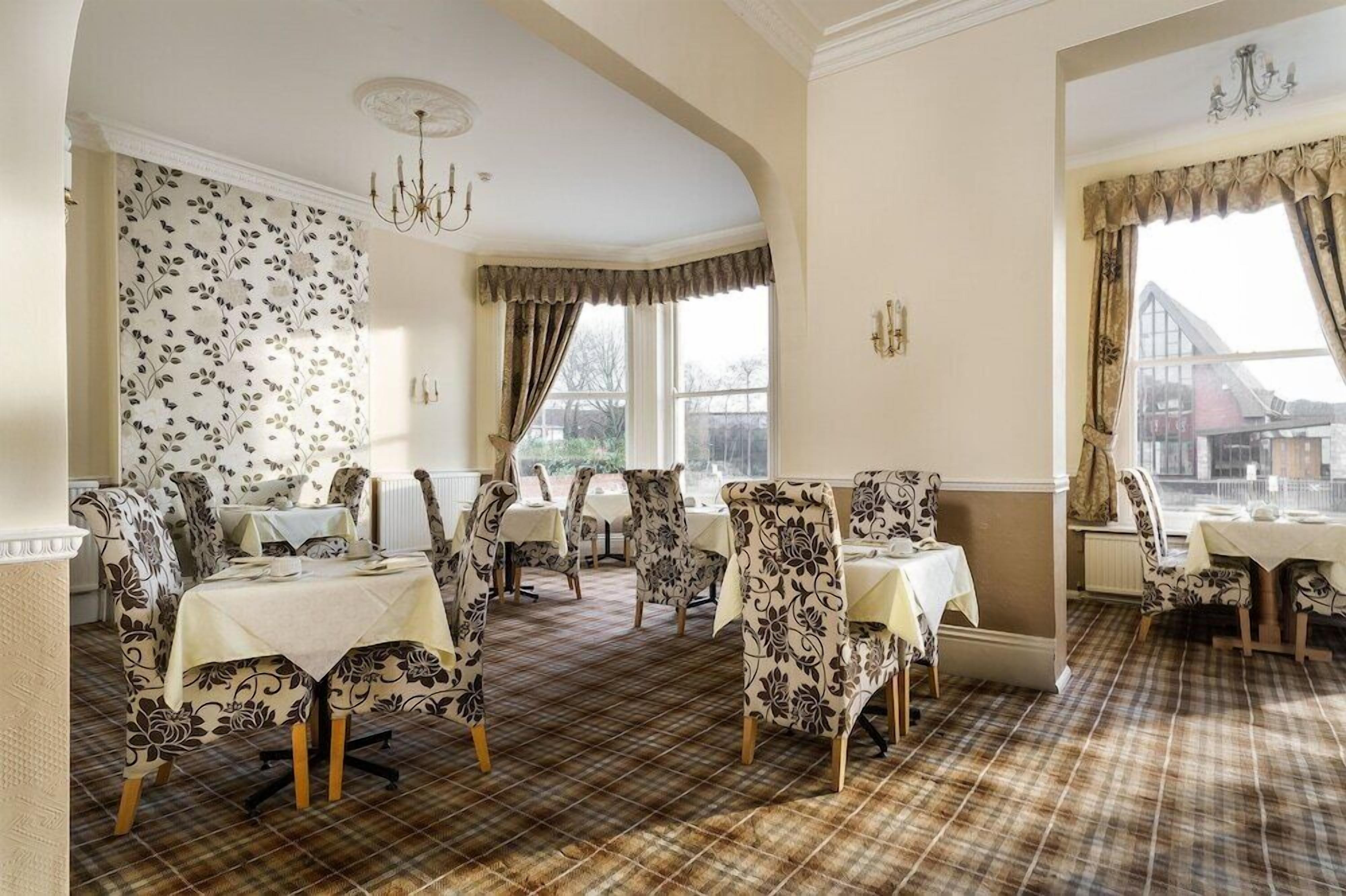 A dining area with patterned chairs and tables, a chandelier, and large windows with curtains.