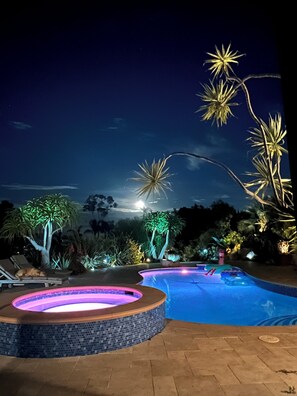 Zona de piscina nocturna a l'aire lliure amb banyera hidromassatge, gandules i plantes il·luminades.