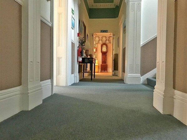 Un pasillo con piso alfombrado, paredes blancas y una puerta que da a una habitación con papel tapiz estampado y un reloj.